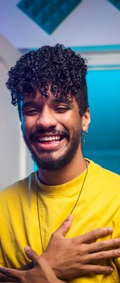 Cheerful young Hispanic blogger crossing hands on chest and smiling while sitting at desk behind smartphone and telling funny story to vlog audience in room with neon illumination at home