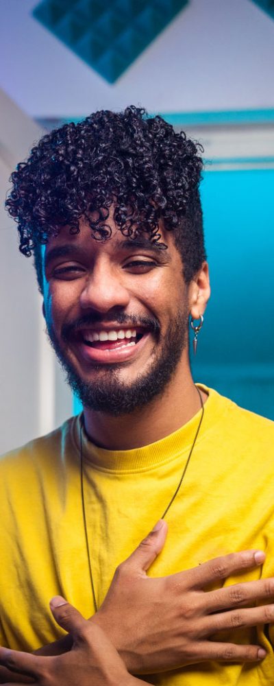 Cheerful young Hispanic blogger crossing hands on chest and smiling while sitting at desk behind smartphone and telling funny story to vlog audience in room with neon illumination at home